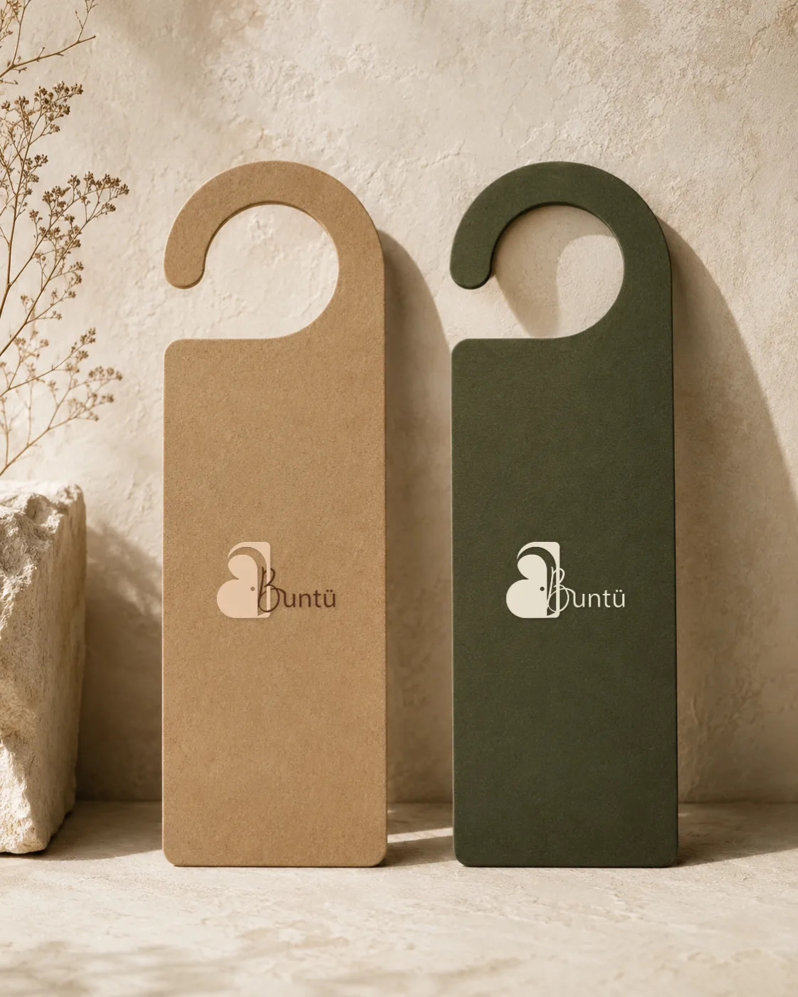 Deux accroche-portes adossés à un mur enduit clair, l'un en kraft, l'autre en olive mat, près d'un bloc de pierre et de fleurs séchées.