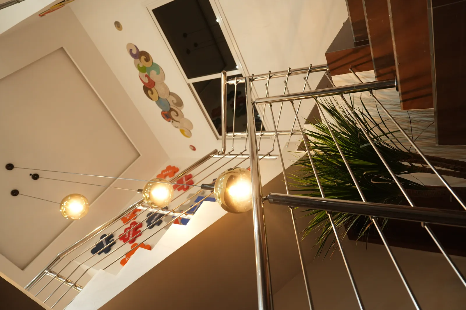 Contre-plongée dans une cage d'escalier : garde-corps chromé à lisses fines, suspensions à globes dorés, murs animés de disques de vannerie colorés et de pièces de puzzle nommant des pays d'Afrique, plante verte.