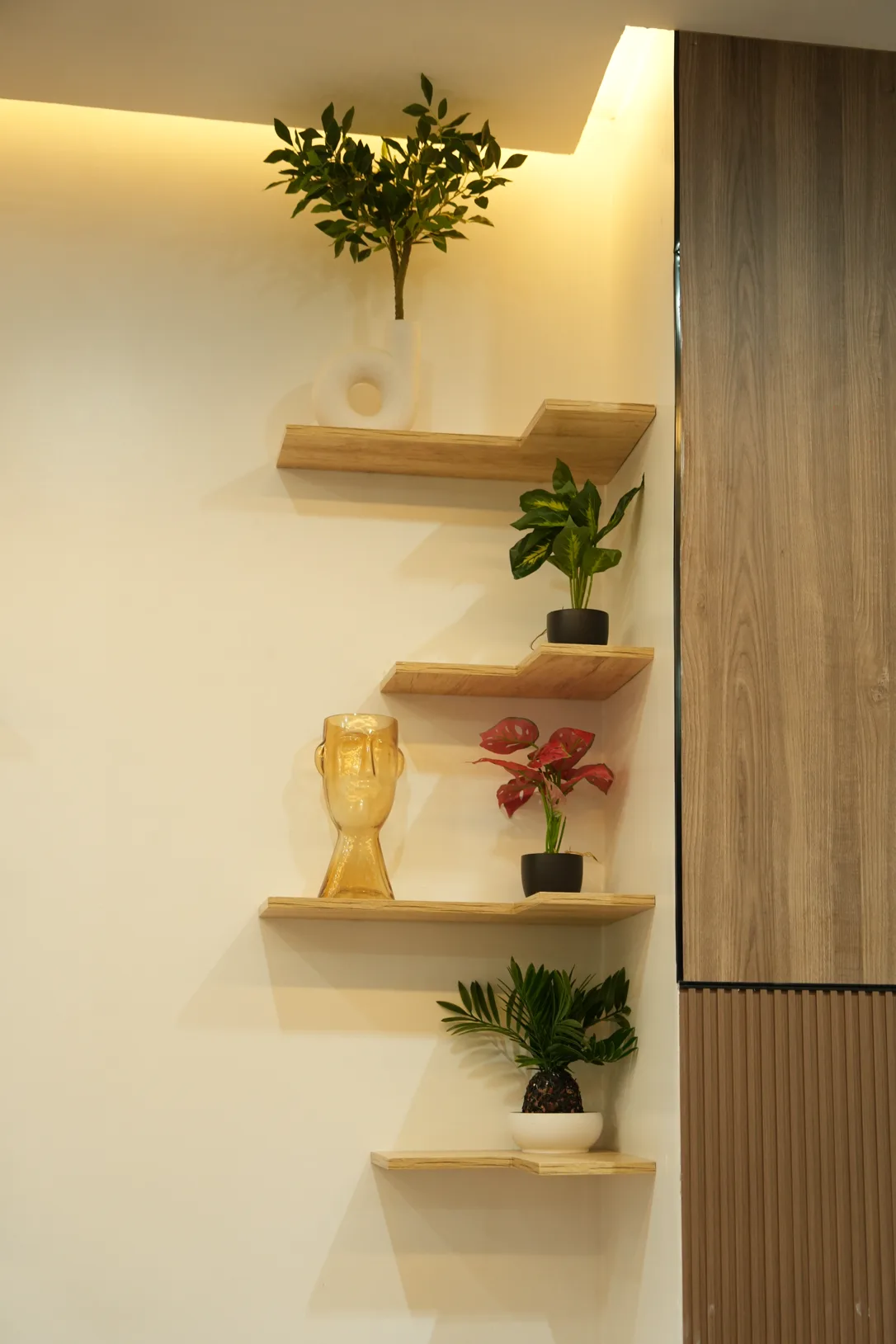 Le même angle réalisé : étagères flottantes en chêne clair posées en quinconce sur le mur blanc, plantes vertes et vases sculpturaux, pan de mur voisin habillé de bois et de tasseaux, éclairage indirect en corniche.