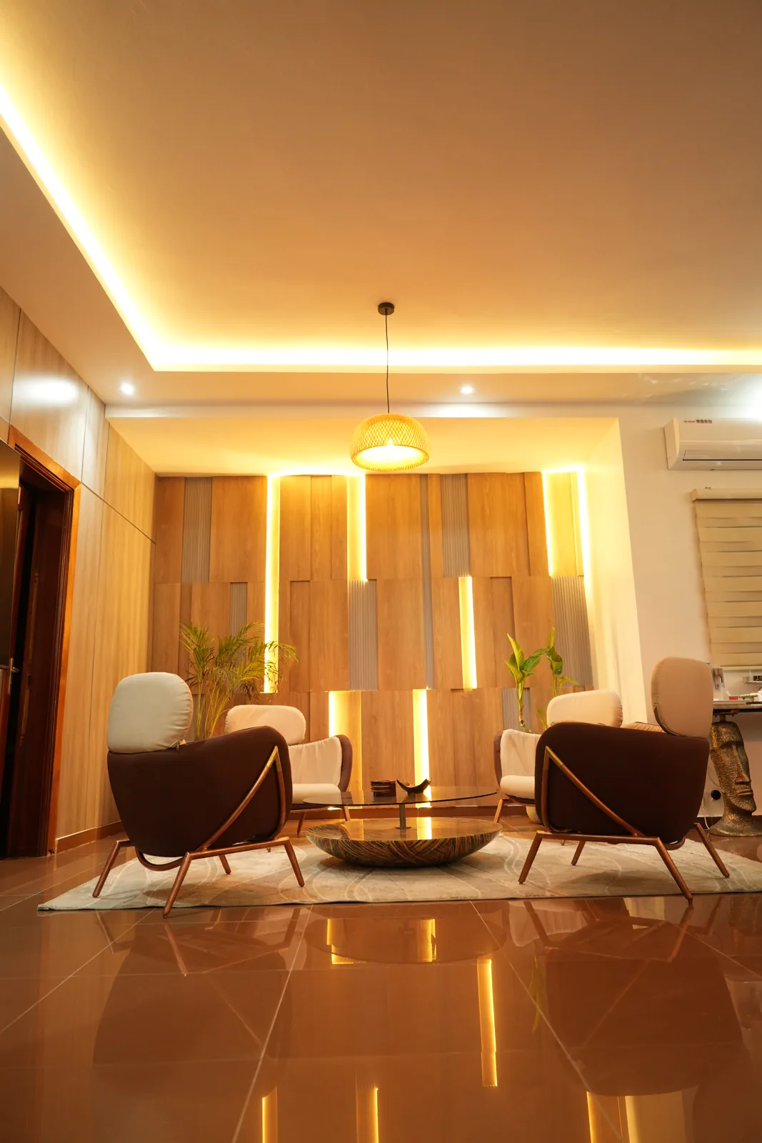 Salon au mur habillé de lames de bois clair entrecoupées de bandeaux lumineux, corniche éclairée, suspension en vannerie, quatre fauteuils crème et brun autour d'une table basse dorée sur un sol poli.