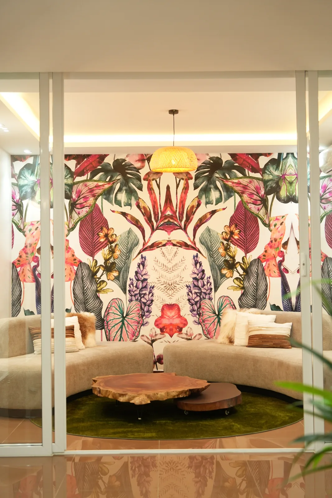 Coin salon vu à travers une verrière blanche : grand papier peint botanique aux feuillages roses, verts et violets, canapé arrondi beige, tables basses en tranches de bois brut sur un tapis vert olive, suspension en vannerie.